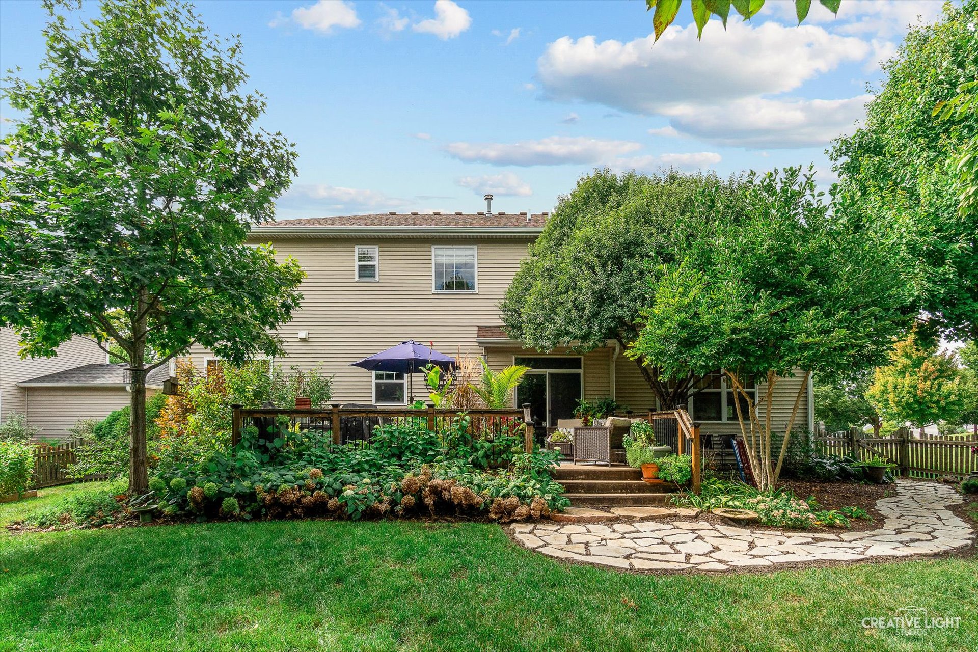 backyard view of a tan house with a flagstone patio, deck and lush landscaping
